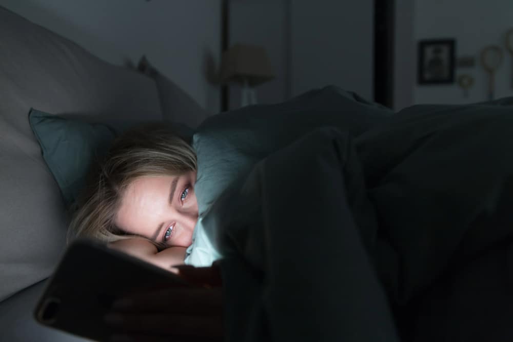 Tired woman lying in bed under the blanket using smartphone at late night as can not sleep.